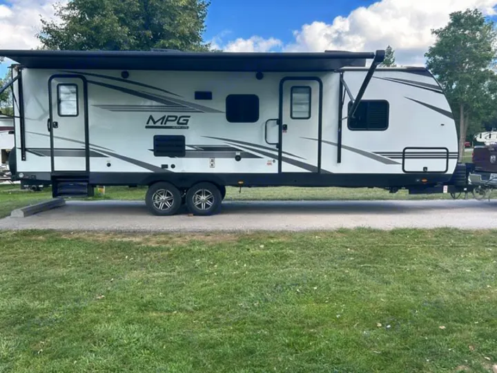 White MPG travel trailer RV rental with awning extended, parked on a paved site in a green, tree-lined campground.. Cruiser RV MPG 2023