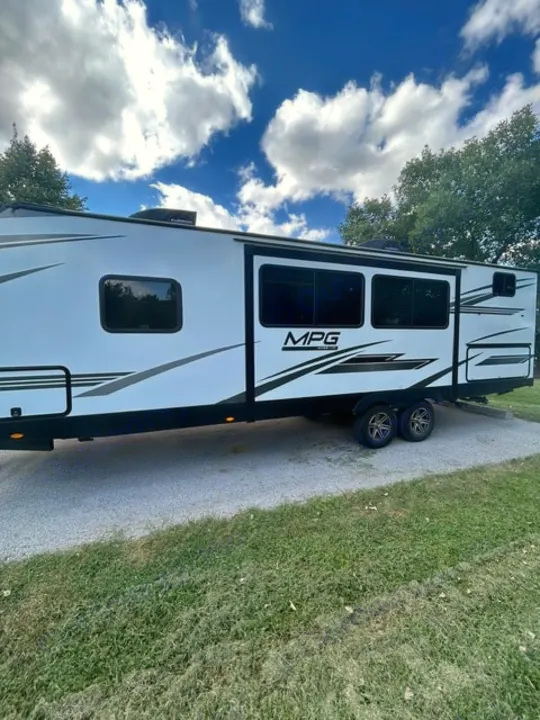 Modern white travel trailer RV rental, parked on gravel with grass, under a bright blue sky with clouds. Ready for camping!. Cruiser RV MPG 2023