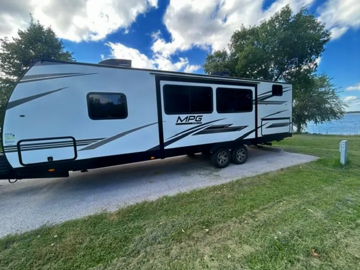 This modern white travel trailer RV rental, model MPG, is parked at a scenic lakefront campsite. Site 49 awaits your camping adventure!. Cruiser RV MPG 2023
