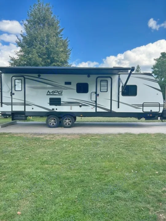 A spacious white MPG travel trailer with an awning extended, parked on asphalt with grass foreground. Great rv rental for camping.. Cruiser RV MPG 2023