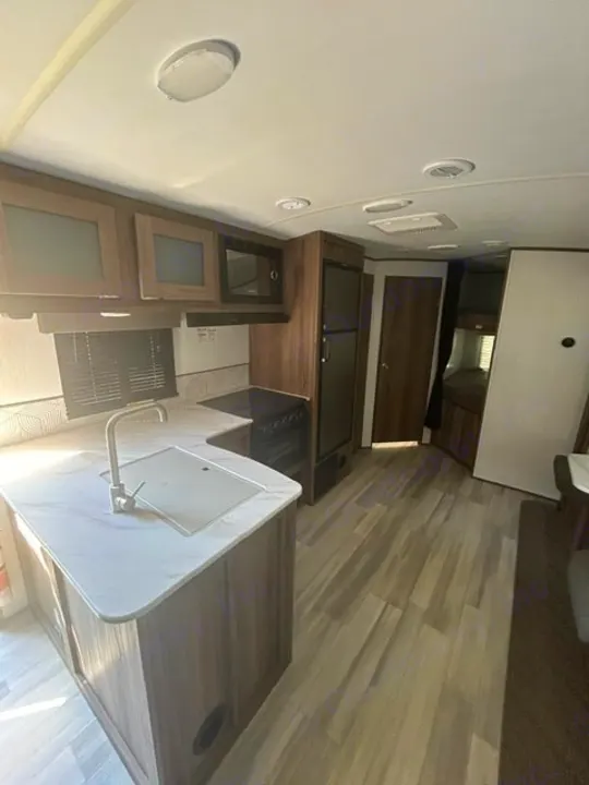 Inside this RV rental, a modern kitchen with wood cabinets, white counters, sink, appliances, and light flooring.. Cruiser RV MPG 2023