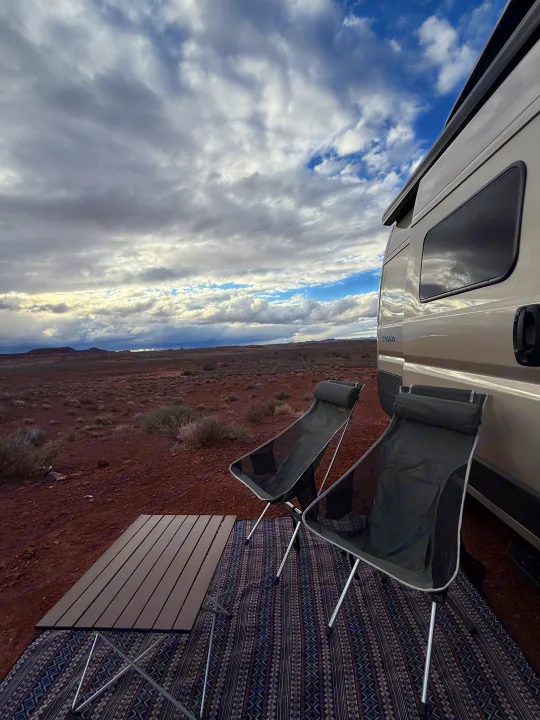 RV rental set up with camping chairs and table on a mat in a scenic desert landscape under a dynamic, cloudy sky.. Custom Camper 2025