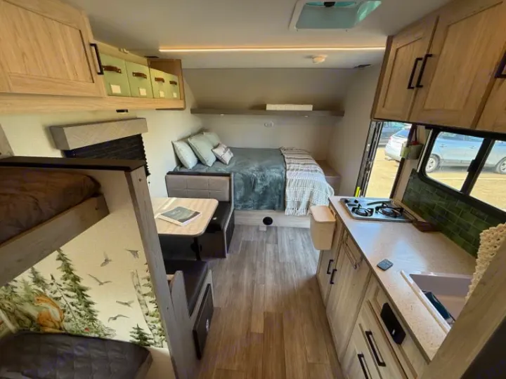 RV rental interior: light wood tones, bunk beds, dining area, kitchen with stove/sink, and a rear bed. Functional camper design.. Heartland RVs Mallard 2025