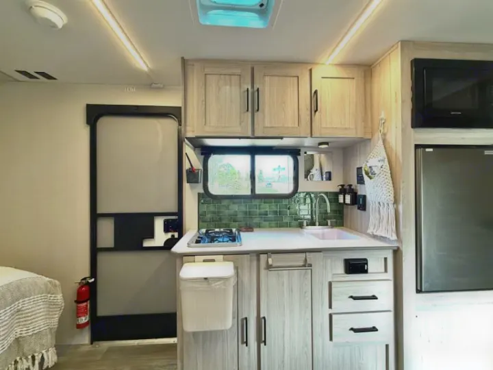Inside an RV rental: a bright kitchen with light wood cabinets, green backsplash, compact stove, sink, fridge, and entry door.. Heartland RVs Mallard 2025