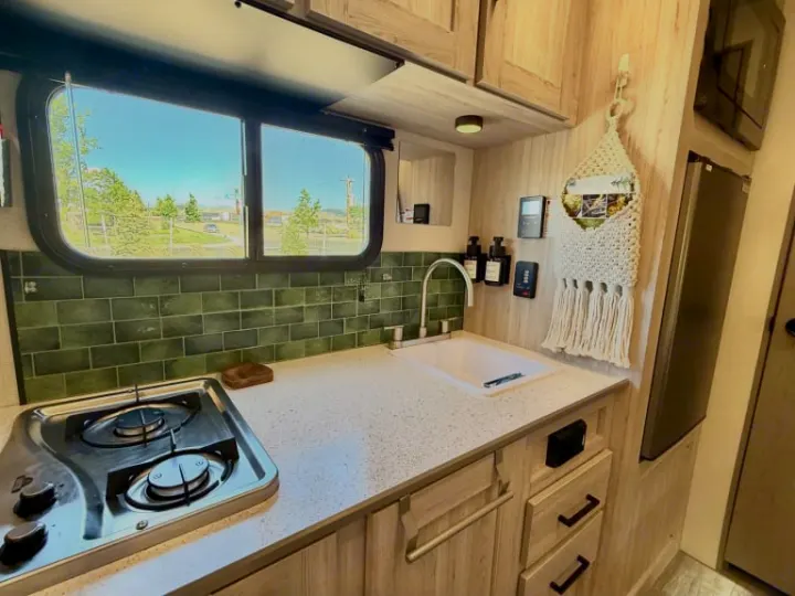 This RV rental kitchen features a green subway tile backsplash, compact stovetop, sink, and cabinets for convenient road trips.. Heartland RVs Mallard 2025