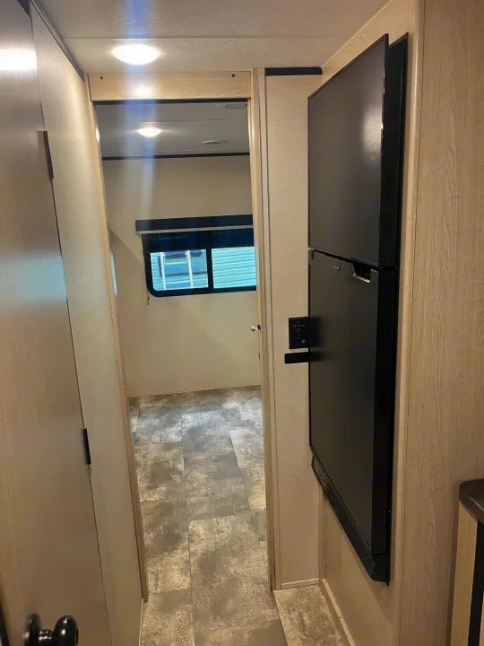 RV rental interior: A narrow hallway with a black refrigerator on the right, light wood walls, and a gray tiled floor.. Coachmen Apex 2023