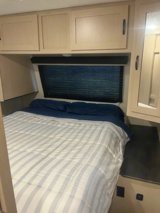 This RV rental bedroom features light wood cabinets, a dark window blind, and a bed with blue sheets and a striped comforter.. Coachmen Apex 2023