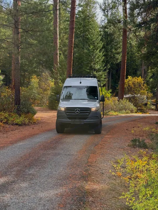 Campervan rv rental, a white Mercedes Sprinter, on a rustic forest road surrounded by tall trees and fall foliage.. Mercedes-Benz Sprinter 2020