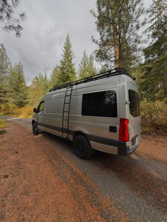 Overcast day with a silver camper van, an ideal **rv rental**, parked on a forest road covered in pine needles.. Mercedes-Benz Sprinter 2020