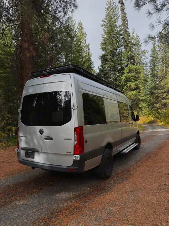Silver Mercedes Sprinter RV rental van parked on a pine-covered forest road, featuring a roof rack for adventure.. Mercedes-Benz Sprinter 2020