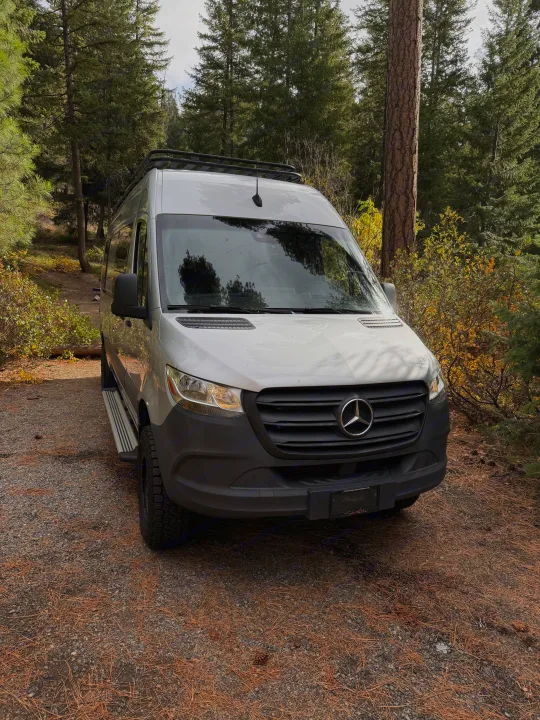 Silver Mercedes Sprinter RV rental parked on a gravel path with pine needles, surrounded by tall evergreen trees.. Mercedes-Benz Sprinter 2020