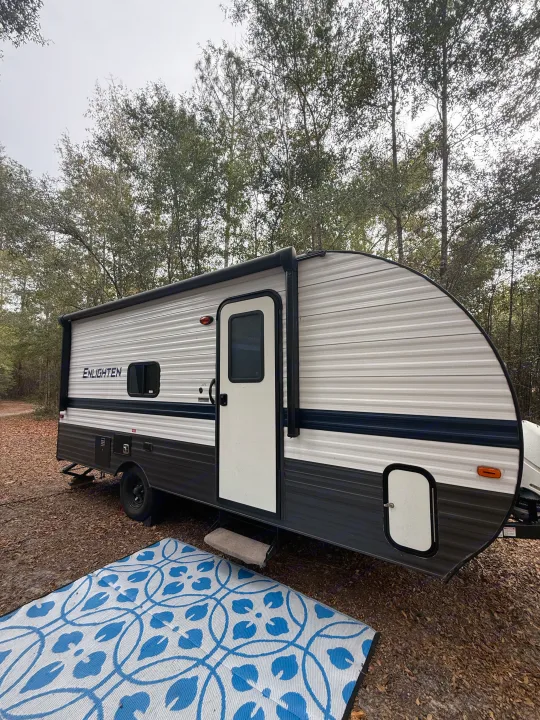 An 'Enlighten' travel trailer RV rental parked in a wooded campsite, featuring a blue outdoor rug and extended awning.. Gulf Stream Enlighten 2021