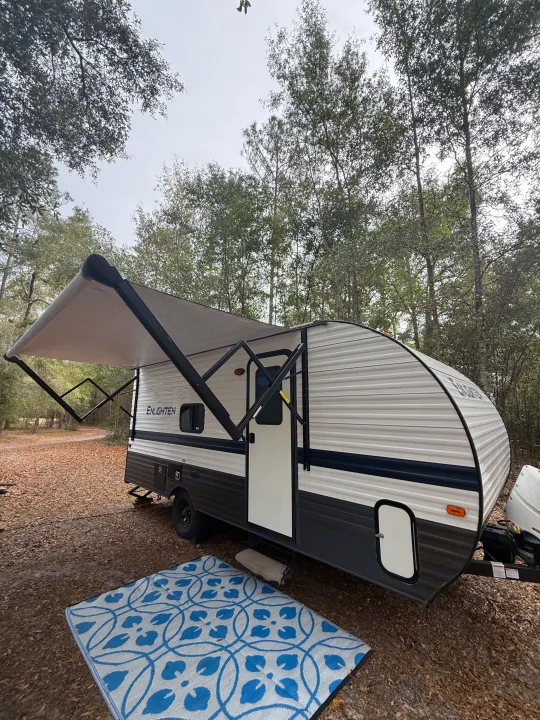 RV rental ready for adventure: a travel trailer with an extended awning and outdoor rug at a wooded campsite.. Gulf Stream Enlighten 2021