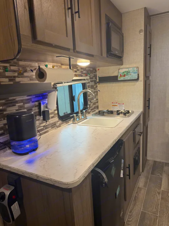 This RV rental galley features a compact kitchen with a sink, stove, microwave, and dehumidifier, plus a "Home of Happy Campers" sign.. Gulf Stream Enlighten 2021