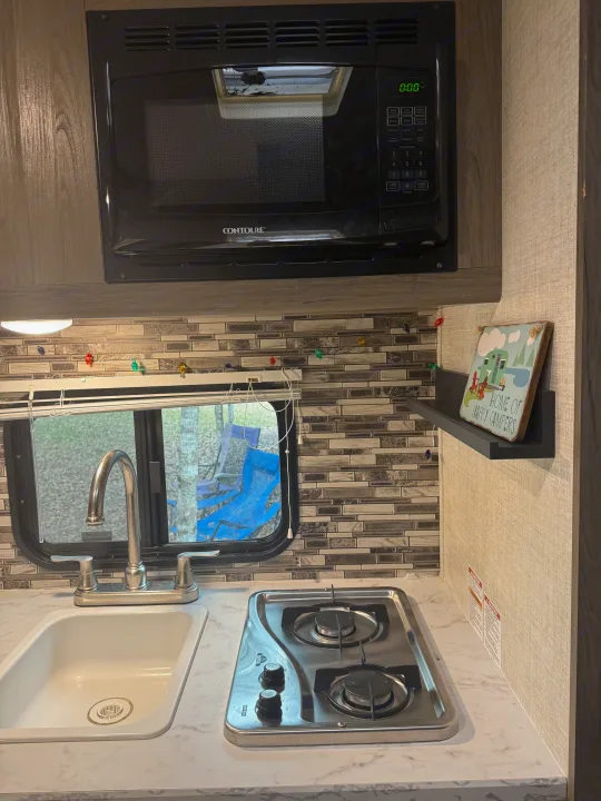 This RV rental kitchen shows a sink, two-burner stovetop, microwave, and a "Home of Happy Campers" sign, ready for travel.. Gulf Stream Enlighten 2021