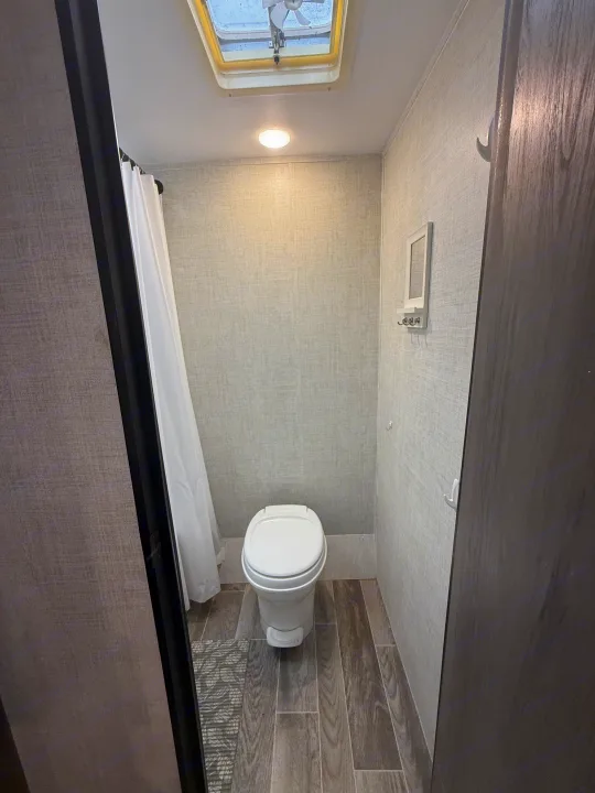 Clean, compact rv rental bathroom featuring a toilet, shower curtain, wood-look floor, roof fan, and integrated lighting.. Gulf Stream Enlighten 2021