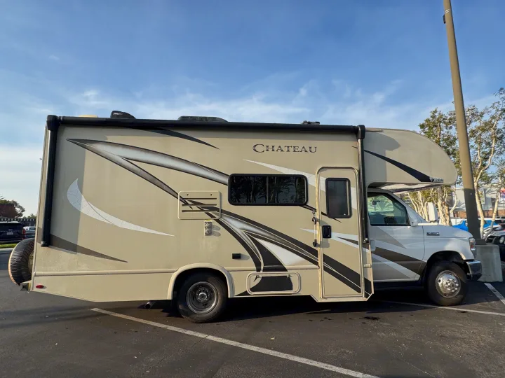 A beige Chateau Class C RV rental with black graphics, parked outdoors under a blue sky. A spare tire is visible at the rear.. Thor Chateau 2019