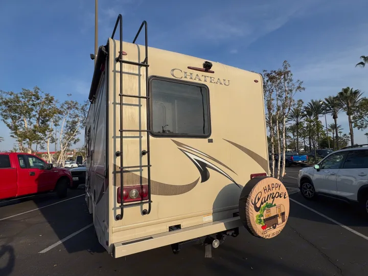 Rear view of a Chateau RV rental in a parking lot, featuring a ladder, window, and a "Happy Camper" spare tire cover.. Thor Chateau 2019