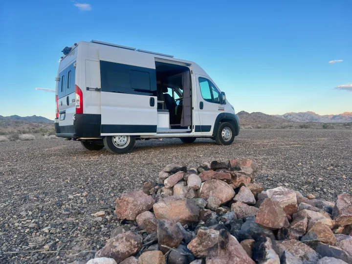 White RV rental van parked in a rocky desert, with mountains under a blue sky and a stone fire pit in the foreground.. Ram Promaster 1500 2020