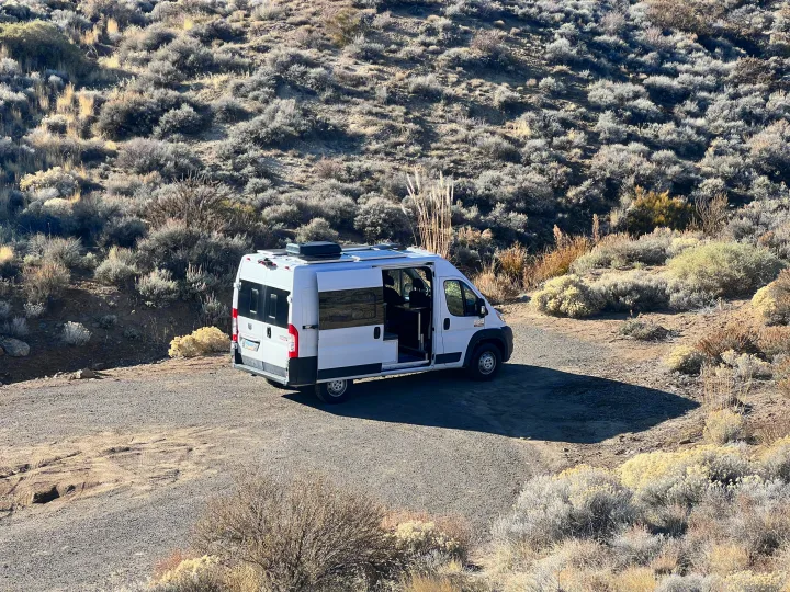 White RV rental camper van parked on a dirt road in a sunny, scrub-covered desert landscape. Side door open for exploring.. Ram Promaster 1500 2020