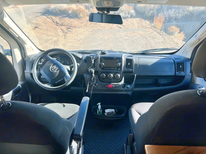 View from an RV rental cabin, showing the dashboard, steering wheel, and front seats, looking out onto a dry, unpaved road.. Ram Promaster 1500 2020