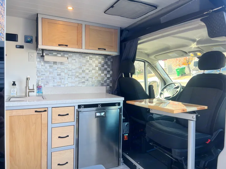 RV rental with a modern camper van kitchen: wood cabinets, tiled backsplash, fridge, sink, and swivel table.. Ram Promaster 1500 2020