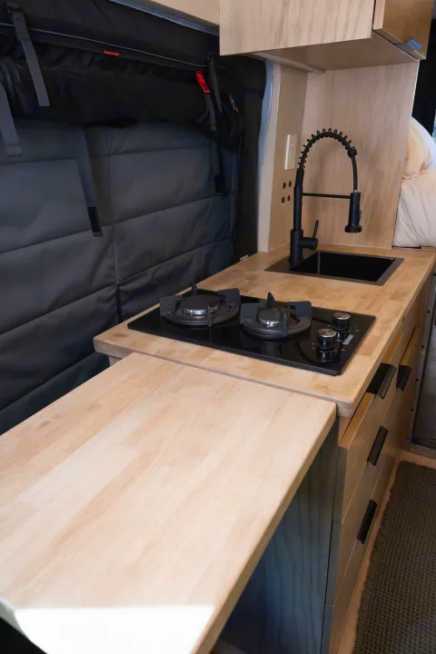 Compact RV rental kitchen features light wood countertops, a black sink, and a built-in gas stove, ready for travel meals.. Ford Transit 2022