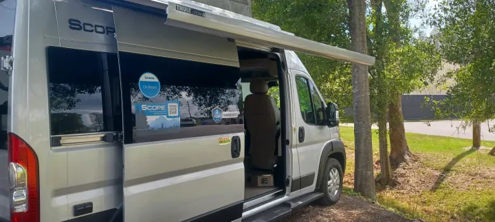 Silver **rv rental** van, branded 'SCOPE', with an extended awning and open side door, parked among trees.. Thor Scope 2023