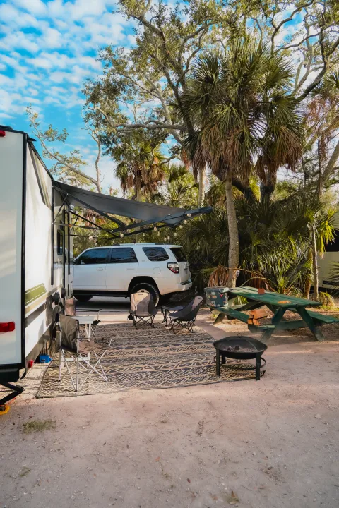 Your RV rental getaway! A spacious campsite with an RV, SUV, awning, chairs, rug, fire pit, and picnic table under trees.. Heartland RVs Eddie Bauer 2025