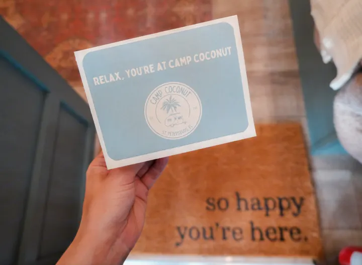 A welcome card for Camp Coconut, featuring an RV logo, held above a "so happy you're here" doormat for this St. Petersburg RV rental.. Heartland RVs Eddie Bauer 2025