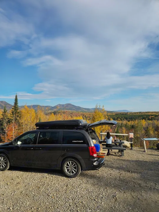 RV rental adventure: Dark minivan with open hatch, roof box, and a person enjoying a scenic mountain view with fall foliage.. Chrysler Grand Caravan 2026