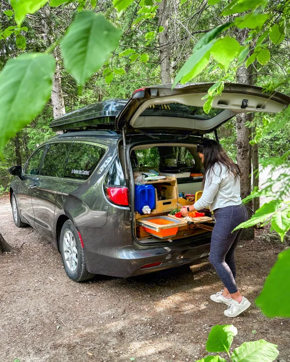 RV rental adventure: Woman preps food at her custom minivan camper kitchen, enjoying a forest campsite.. Chrysler Grand Caravan 2022