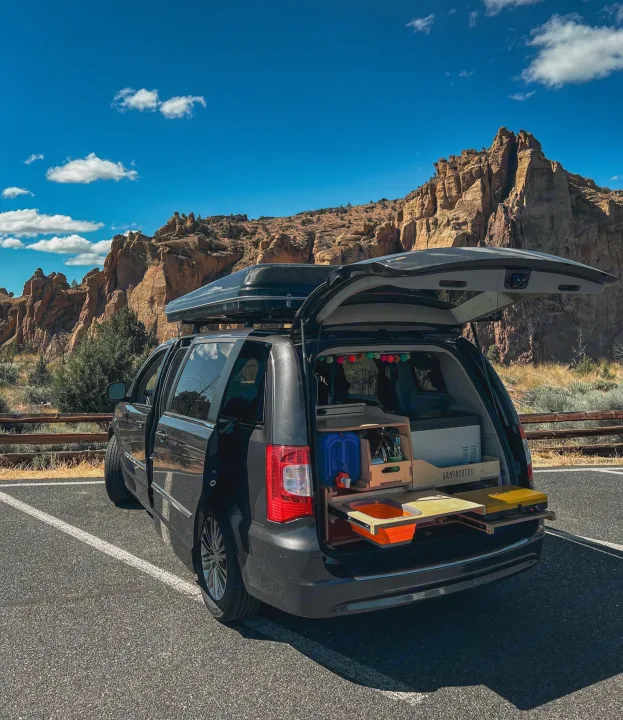 RV rental minivan with custom tailgate kitchen & roof box, ideal for a van life road trip. Scenic rock formations in the background.. Chrysler Grand Caravan 2025