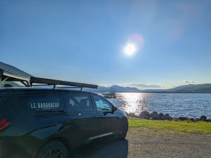 Explore nature with an rv rental! Black "Le Baroudeur" van by a sun-drenched lake and mountains. Ideal for van life travel.. Chrysler Pacifica (hydride) 2022
