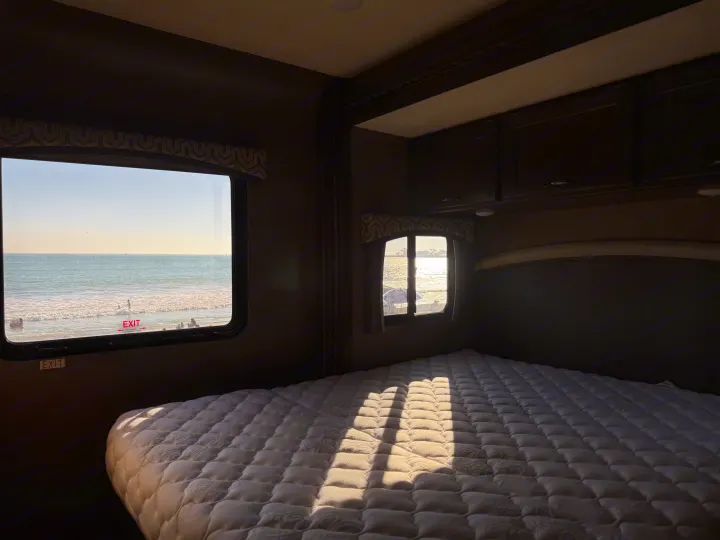 Bright morning in an RV rental bedroom overlooking the ocean. Sunbeams highlight the quilted bed, with beachgoers outside.. Thor Chateau 2019