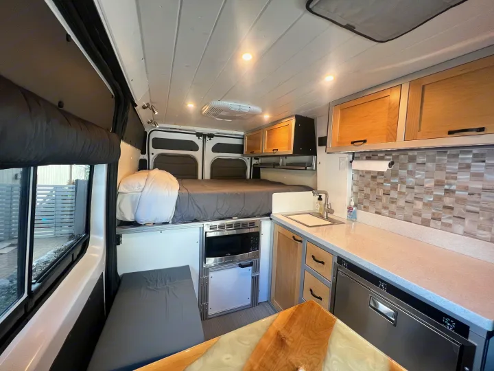 This RV rental interior showcases a cozy bed, compact kitchen with sink & microwave, and a dining area. Ready for your adventure.. Ram Promaster 1500 2020