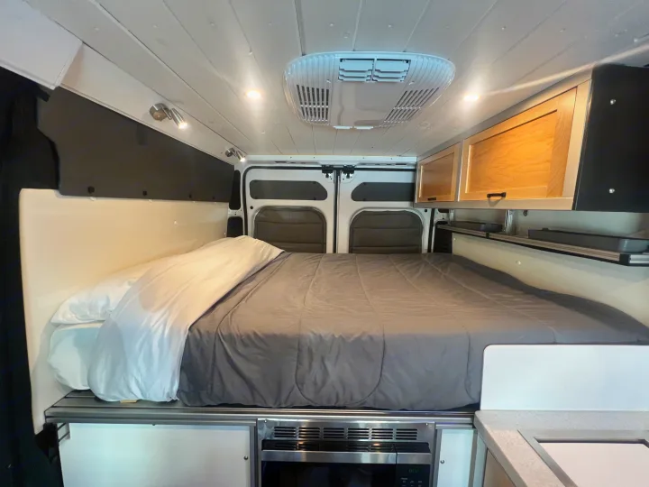 This cozy RV rental interior features a queen-sized bed, overhead storage cabinets, and a convenient under-bed microwave.. Ram Promaster 1500 2020