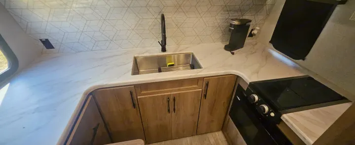 Compact **rv rental** kitchen featuring a white marble-look countertop, stainless sink, black faucet, wooden cabinets, Keurig, and stove.. Grand Design Imagine AIM 2023