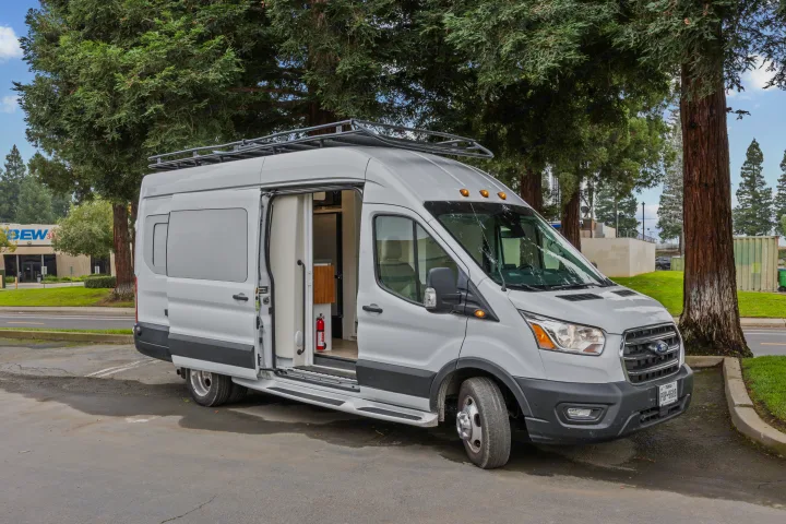 White Ford Transit RV rental parked on asphalt, door open to reveal a built-in kitchen, storage, and a red fire extinguisher.. Coachmen Beyond 2021