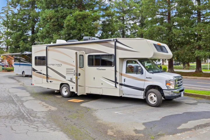 Beige and brown Coachmen Leprechaun rv rental parked with door open on an asphalt lot, tall trees behind.. Coachmen Leprechaun 2019