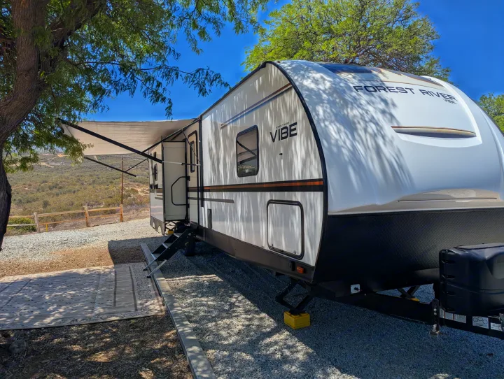 A white Forest River Vibe RV rental is parked at a shaded campsite with its awning extended over a gravel site, ready for use.. Forest River Vibe 2020