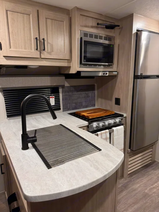 This modern RV rental kitchen features light wood cabinets, stainless appliances, a sink, and a stove. Perfect for travel.. Forest River Aurora Aurora 2024