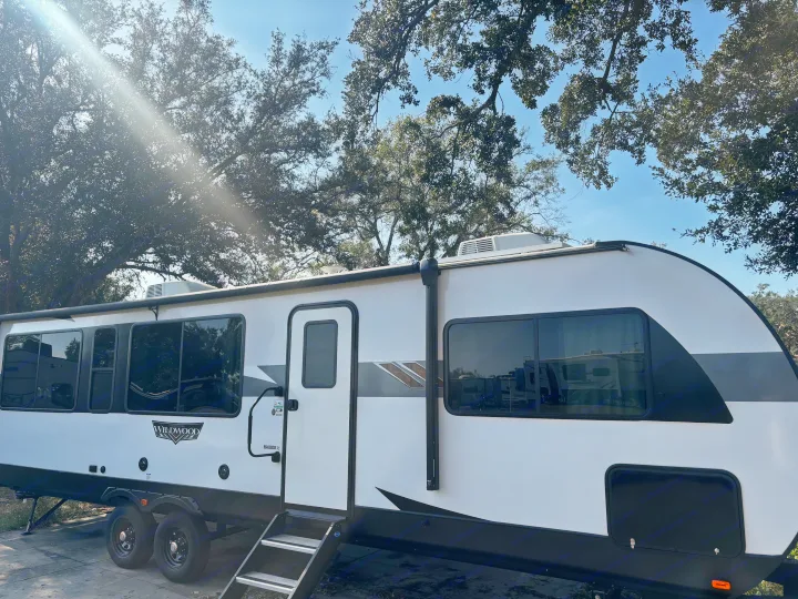Bright white Wildwood RV rental parked under dappled sunlight through trees. A modern travel trailer ready for your outdoor adventure.. Forest River Wildwood Heritage Glen 2024