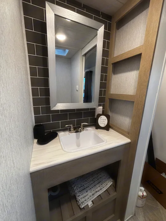 This RV rental bathroom showcases a functional vanity with a sink, mirror, black subway tile, and open storage for essentials.. Forest River Wildwood Heritage Glen 2024