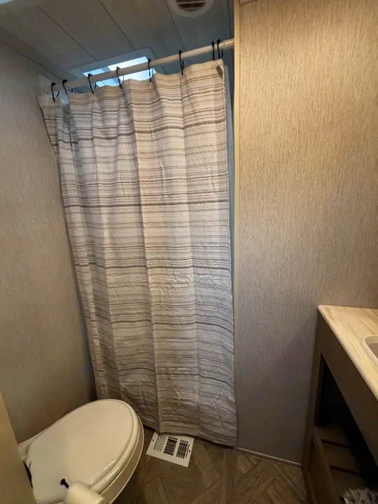 RV rental's compact bathroom showing a white toilet, a grey/white striped shower curtain, and a small vanity to the right.. Forest River Wildwood Heritage Glen 2024