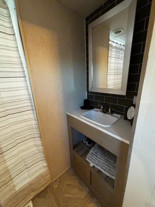 RV rental bathroom with a vanity, white sink, mirror, black subway tile backsplash, and a striped shower curtain to the left.. Forest River Wildwood Heritage Glen 2024