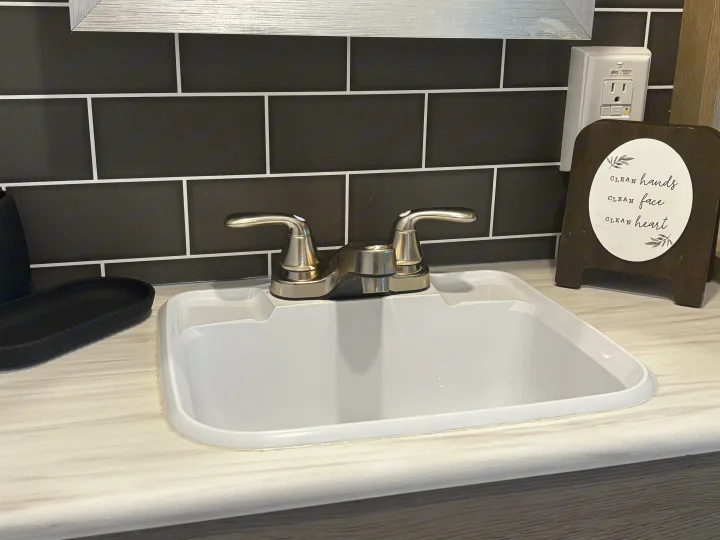 This RV rental bathroom features a compact sink, chrome faucet, black subway tile backsplash, GFCI outlet, and a decorative sign.. Forest River Wildwood Heritage Glen 2024