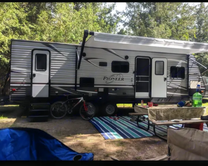 Relaxing at a shady campsite in an RV rental, featuring an extended awning, bicycle, and picnic area for outdoor comfort.. Pioneer Bunkhouse 2019