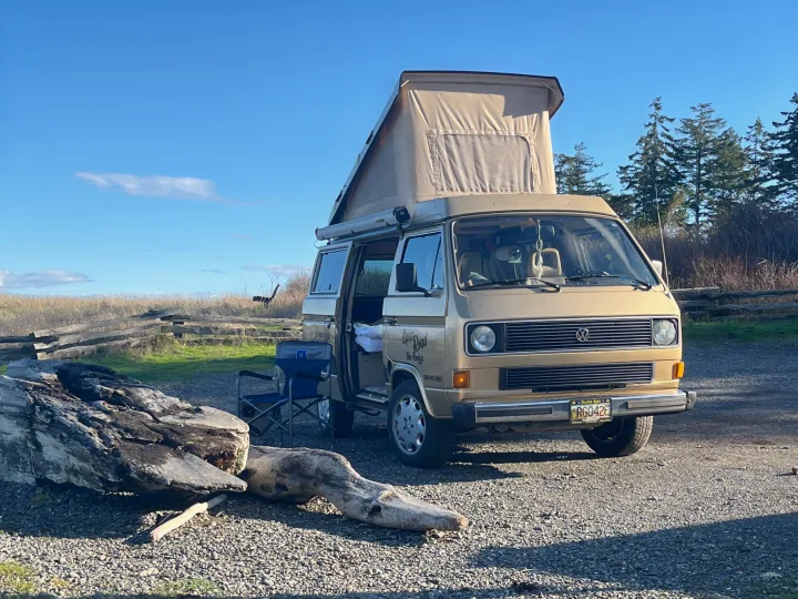 A beige VW camper van (rv rental) from Open Road, pop-top extended, parked on gravel with driftwood and nature around.. Volkswagen T3 Westfalia 1985