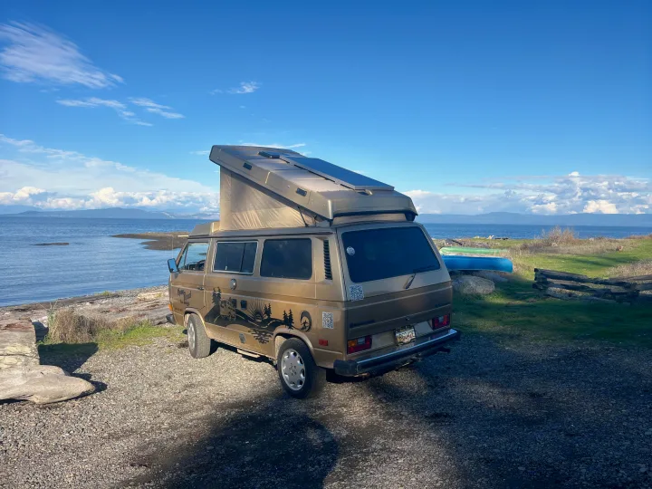 Open Road RV rental: Golden VW camper van with solar panels parked by the ocean, mountains, and blue sky.. Volkswagen T3 Westfalia 1985